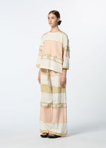 A person in a studio wears HaaT's KASANEGAKE jacquard-woven top with three-quarter sleeves and wide-leg pants, both featuring pastel pink, white, and beige horizontal stripes. They sport slicked-back hair and black sandals.