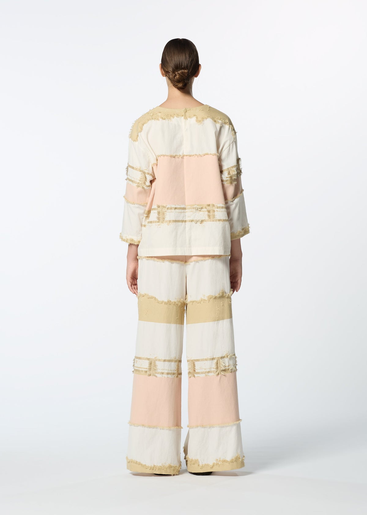 A person stands facing away in the HaaT KASANEGAKE jacquard woven set, featuring a layered top and wide-leg pants with beige, cream, and light pink color blocks and fringe accents. The background is plain white. view 7 of 7