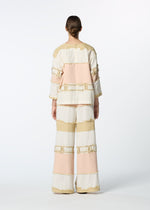 A person stands facing away in the HaaT KASANEGAKE jacquard woven set, featuring a layered top and wide-leg pants with beige, cream, and light pink color blocks and fringe accents. The background is plain white.
