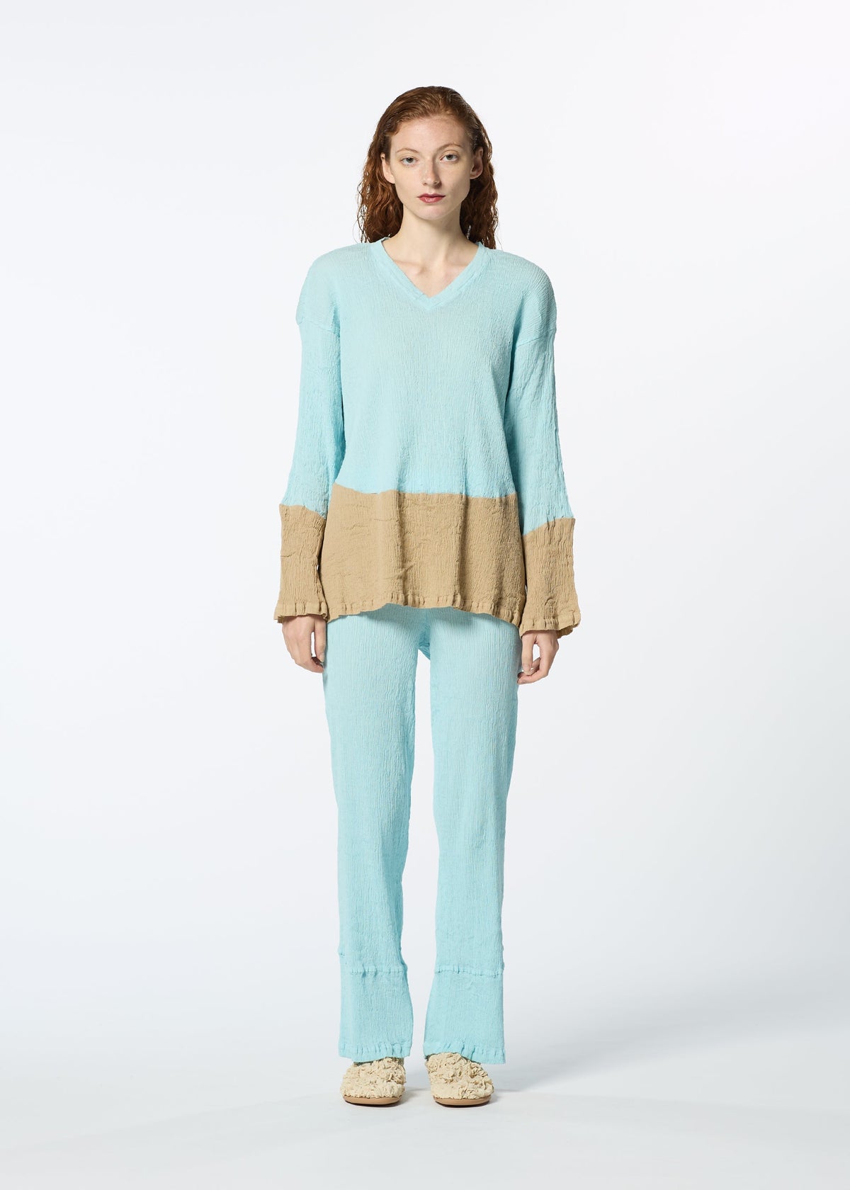 A person with long, wavy hair models the HaaT KYO CHIJIMI STREAM: a loose, lightweight textured top in light blue with beige sleeve and waist accents, standing against a white background. view 4 of 6