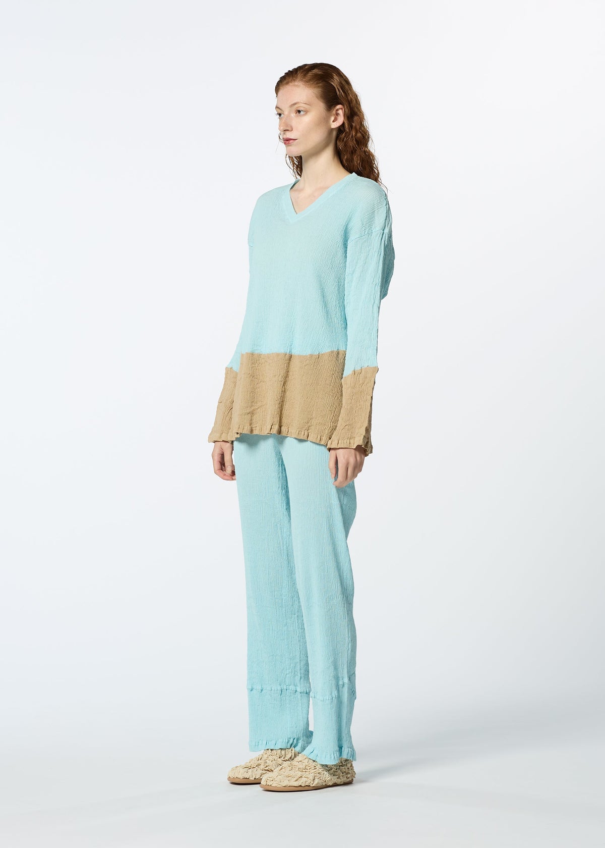 A person faces left, wearing HaaT's KYO CHIJIMI STREAM: a loose, light blue long-sleeved top with tan blocks on the sleeves and midsection, matching blue pants, and beige slippers, against a plain white background. view 5 of 6
