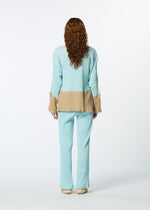 A person with long curly hair stands facing away, wearing HaaT’s KYO CHIJIMI STREAM—a loose, light blue long-sleeved top with tan cuffs and a matching back band—plus beige shoes, all against a plain white background.
