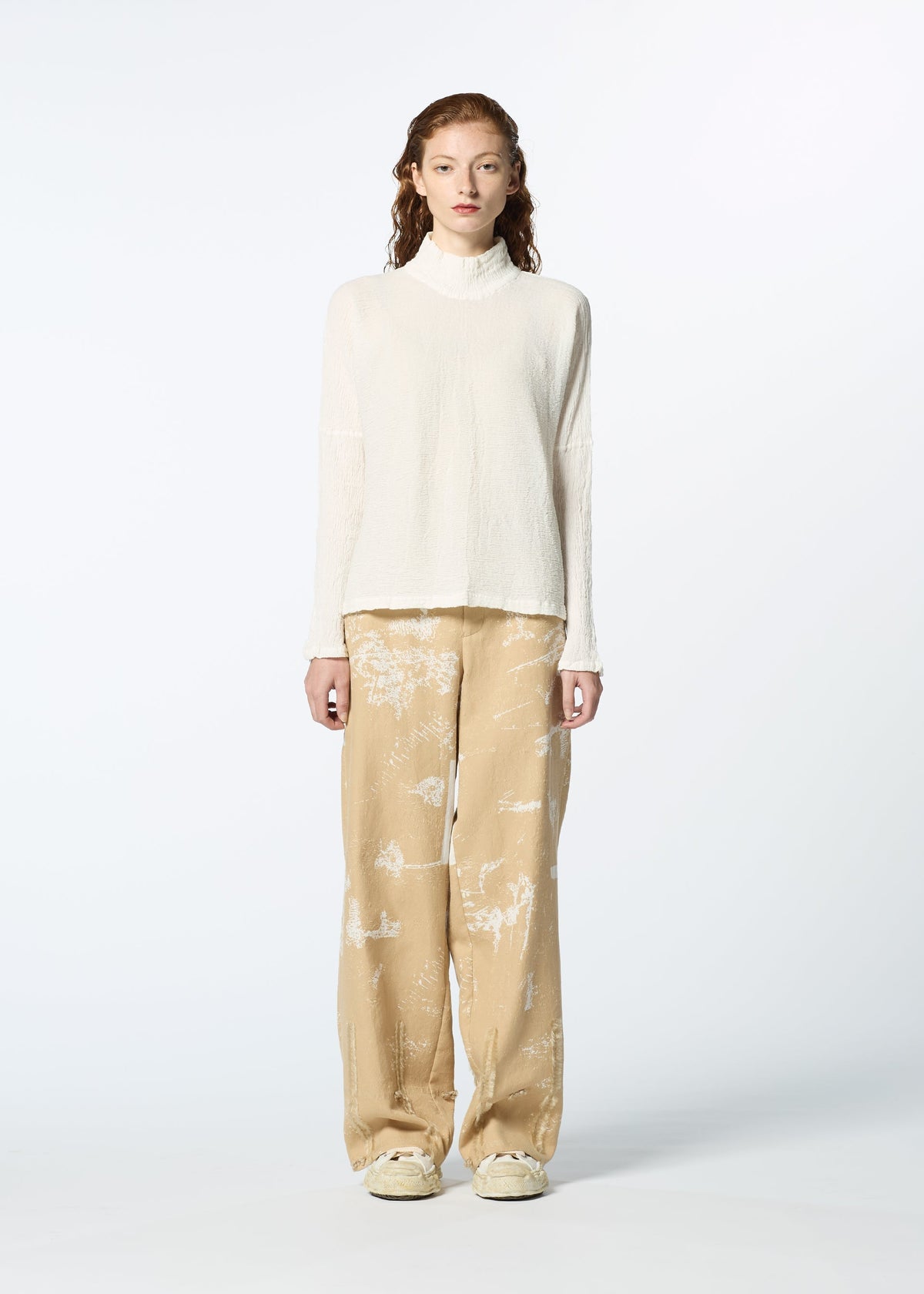 A person faces forward, wearing HaaT's KYO CHIJIMI 2WAY JANUARY 1 high-neck white sweater, beige wide-leg pants with a white floral pattern, and white shoes against a plain light background. view 5 of 7