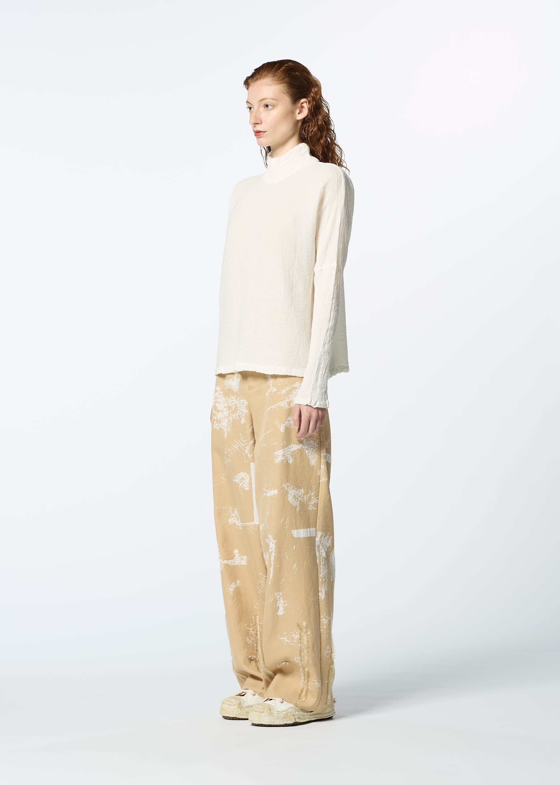 Zoomed view A person with long, wavy hair stands sideways against a plain light background, wearing HaaT's KYO CHIJIMI 2WAY JANUARY 1 high-neck sweater and wide beige patterned pants, paired with cream sneakers.