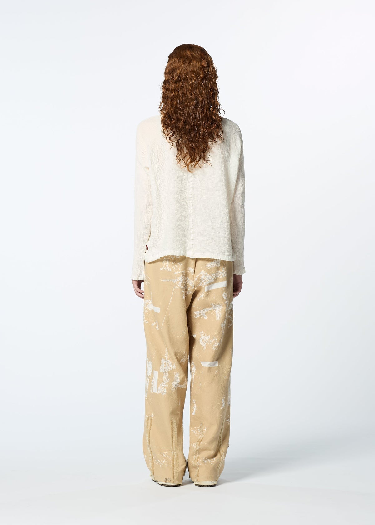 A person with long, curly hair is seen from behind wearing the HaaT KYO CHIJIMI 2WAY JANUARY 1 high-neck, cream top and loose beige pants with white abstract patterns, set against a plain white background. view 7 of 7