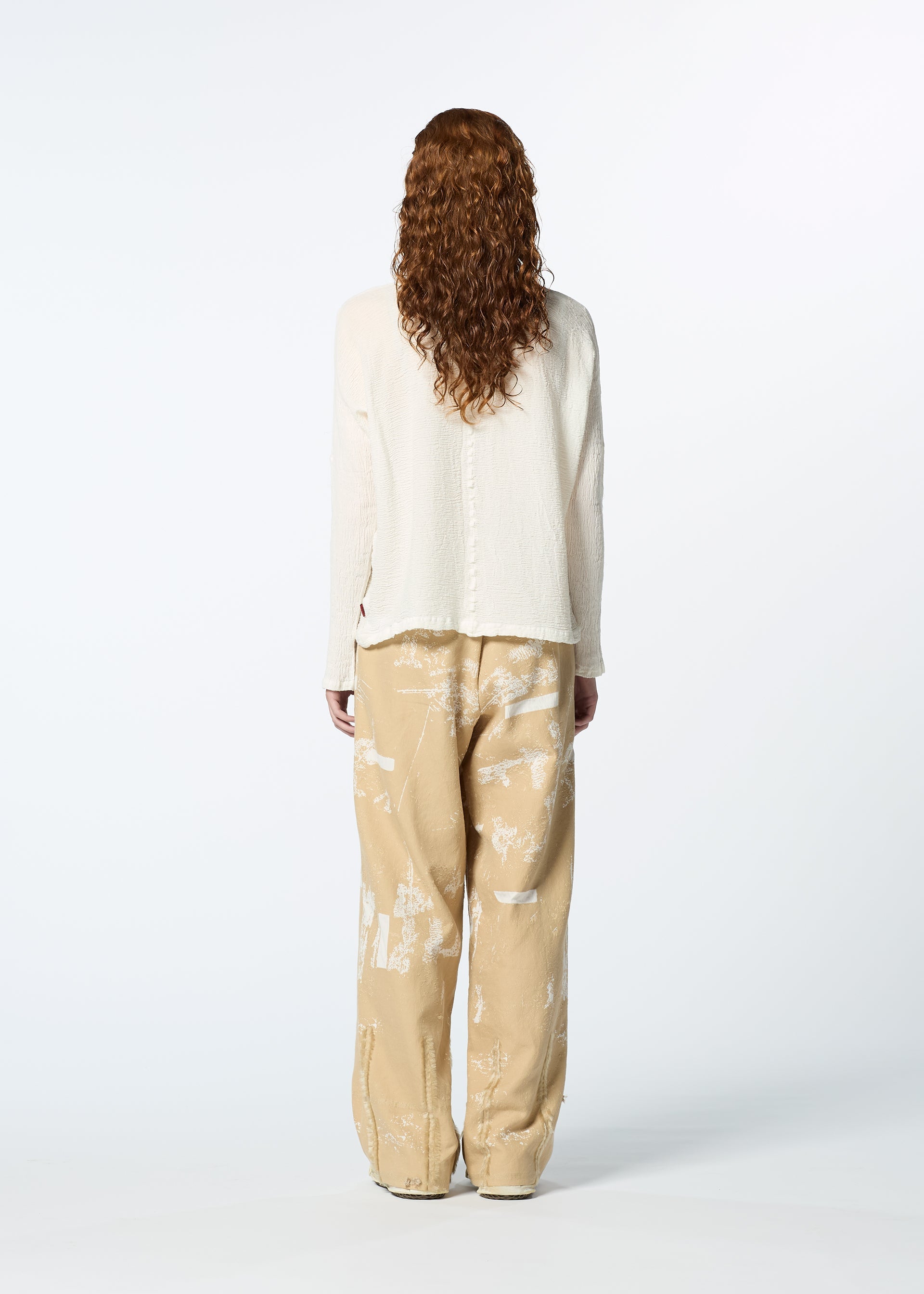 Zoomed view A person with long, curly hair is seen from behind wearing the HaaT KYO CHIJIMI 2WAY JANUARY 1 high-neck, cream top and loose beige pants with white abstract patterns, set against a plain white background.