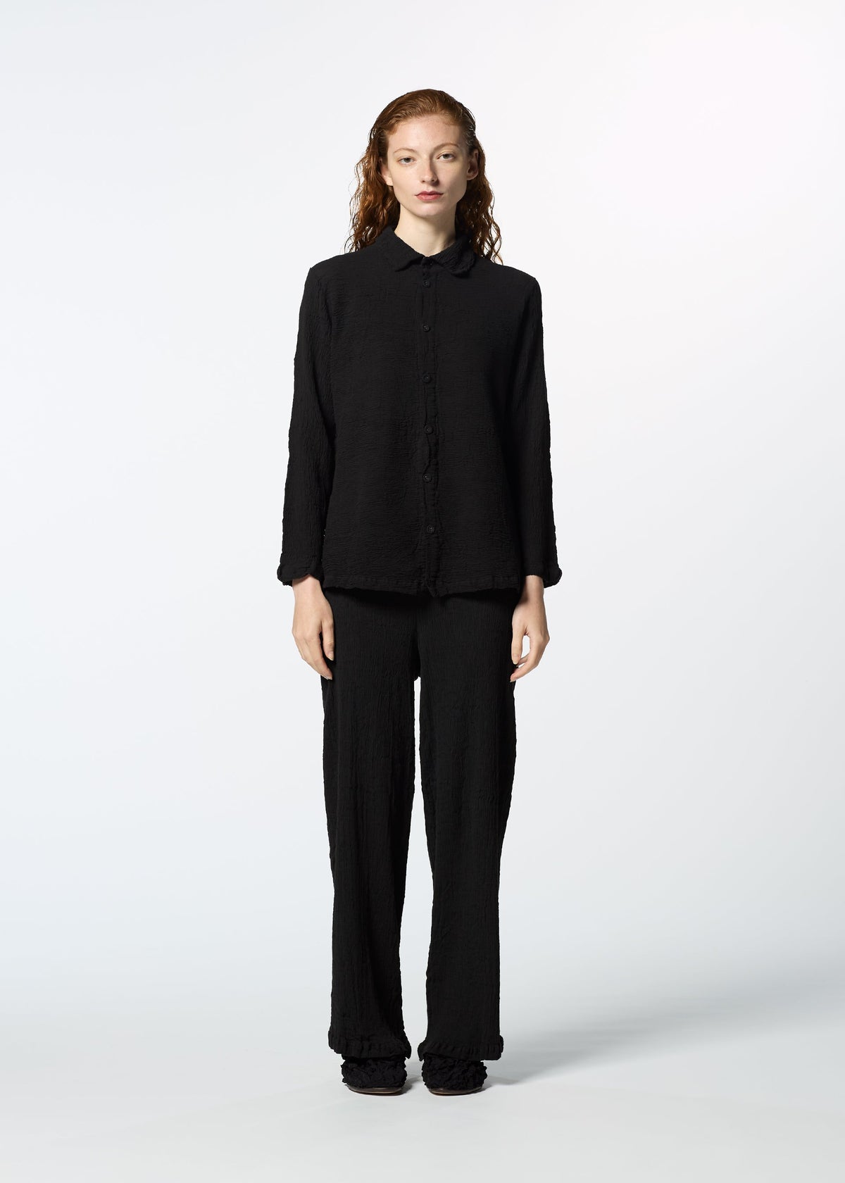 A person with long, wavy hair stands against a light background, wearing the HaaT KYO CHIJIMI 2WAY JANUARY 1 long-sleeved shirt and matching black pants with black shoes. view 5 of 8