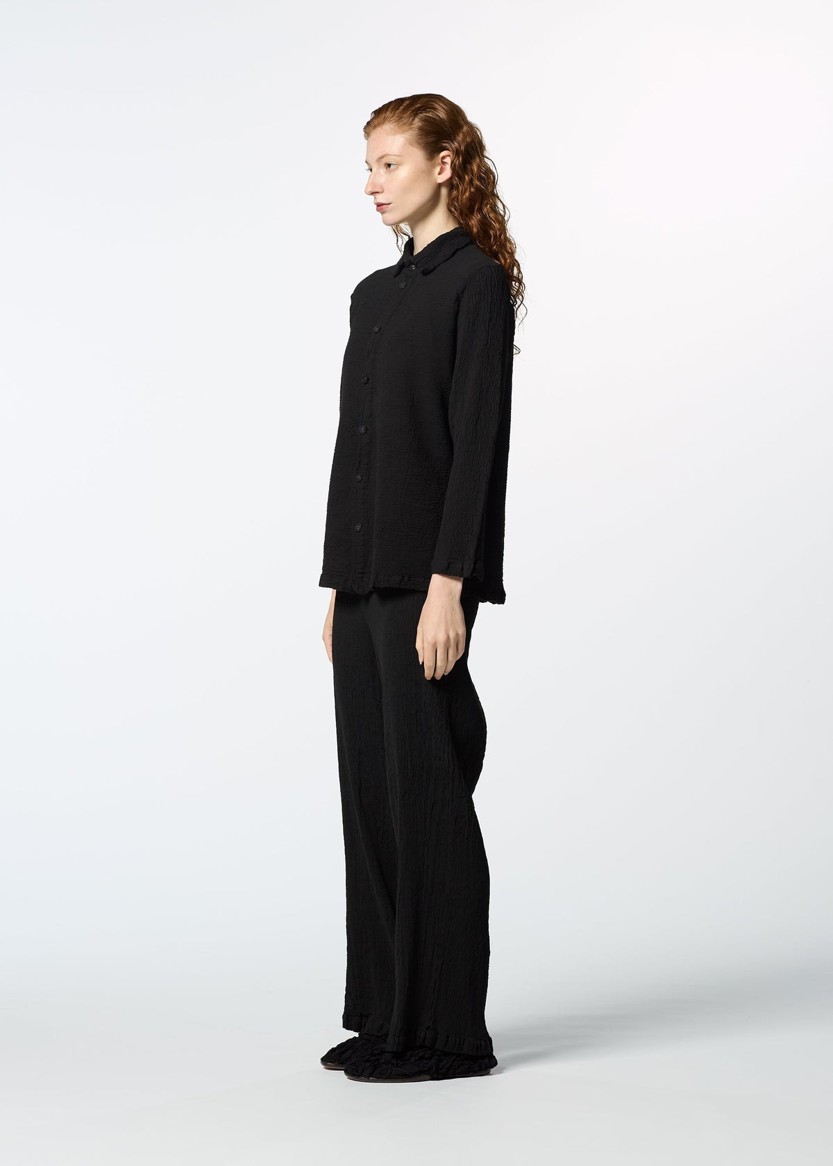 A person with long red hair wears HaaT’s KYO CHIJIMI 2WAY JANUARY 1 set— a loose black long-sleeved shirt and matching wide-leg pants—standing in profile against a plain white background. view 6 of 8