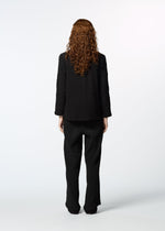 A person with long, curly red hair stands facing away against a white background, wearing the HaaT KYO CHIJIMI 2WAY JANUARY 1: a loose black suit with straight-leg pants and garment-dyed blazer over a long-sleeved shirt.