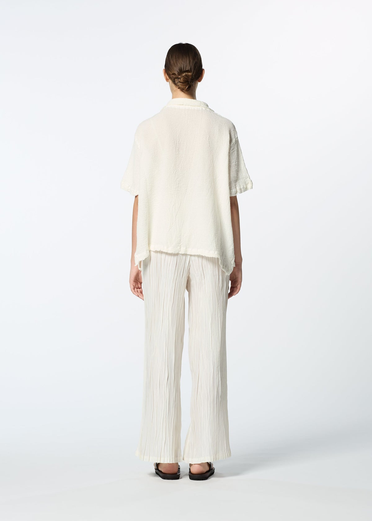 A person faces away, dressed in the HaaT KYO CHIJIMI 2WAY JUNE 1 short-sleeved shirt and wide, pleated, off-white pants with black sandals against a plain white background. view 7 of 7