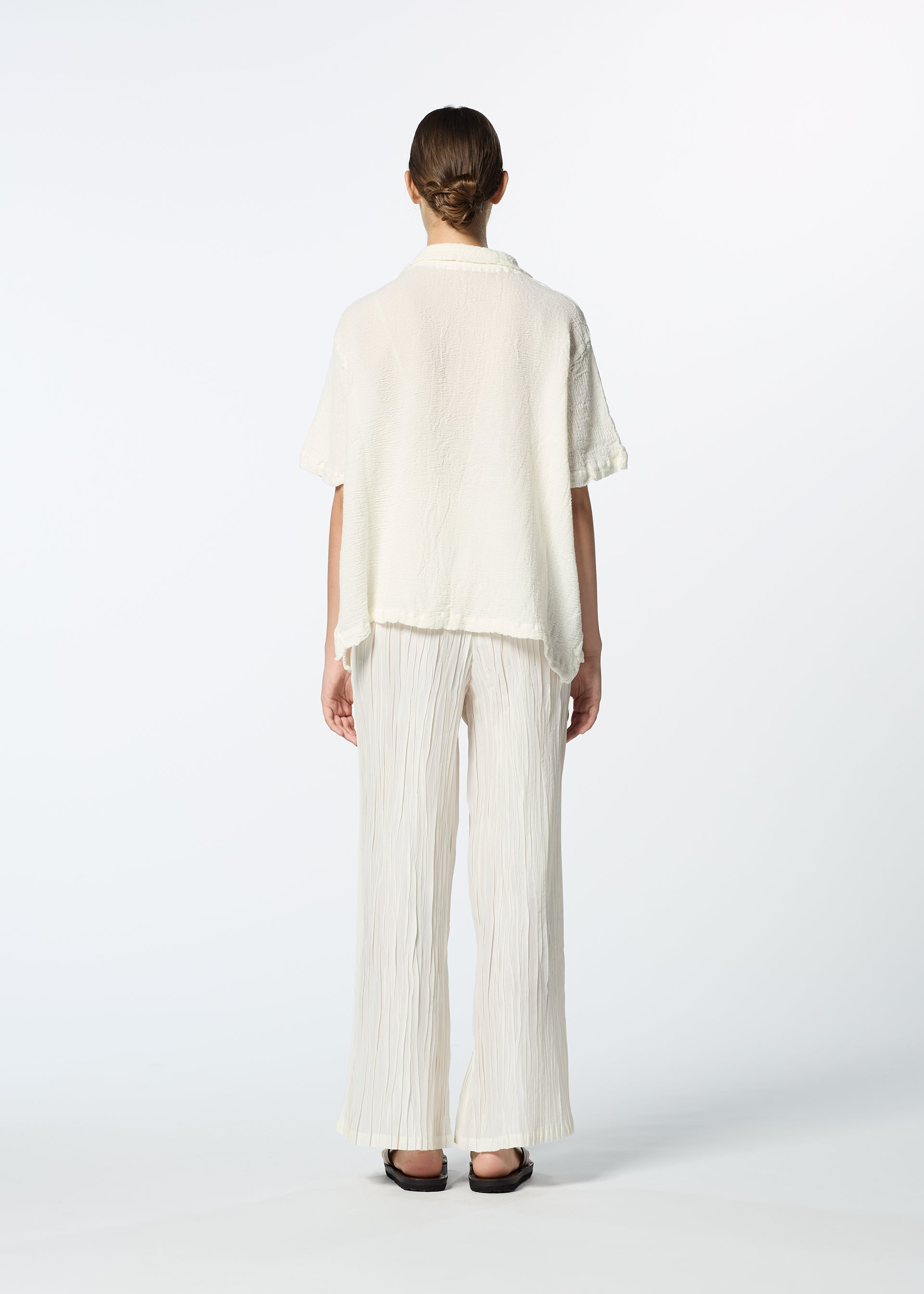 Zoomed view A person faces away, dressed in the HaaT KYO CHIJIMI 2WAY JUNE 1 short-sleeved shirt and wide, pleated, off-white pants with black sandals against a plain white background.