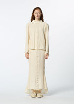 A woman with long, wavy hair wears HaaT's TENMOKU COTTON cream-colored, garment-dyed long-sleeved top and matching ankle-length skirt with subtle front ruffles, paired with beige shoes, standing against a white background.