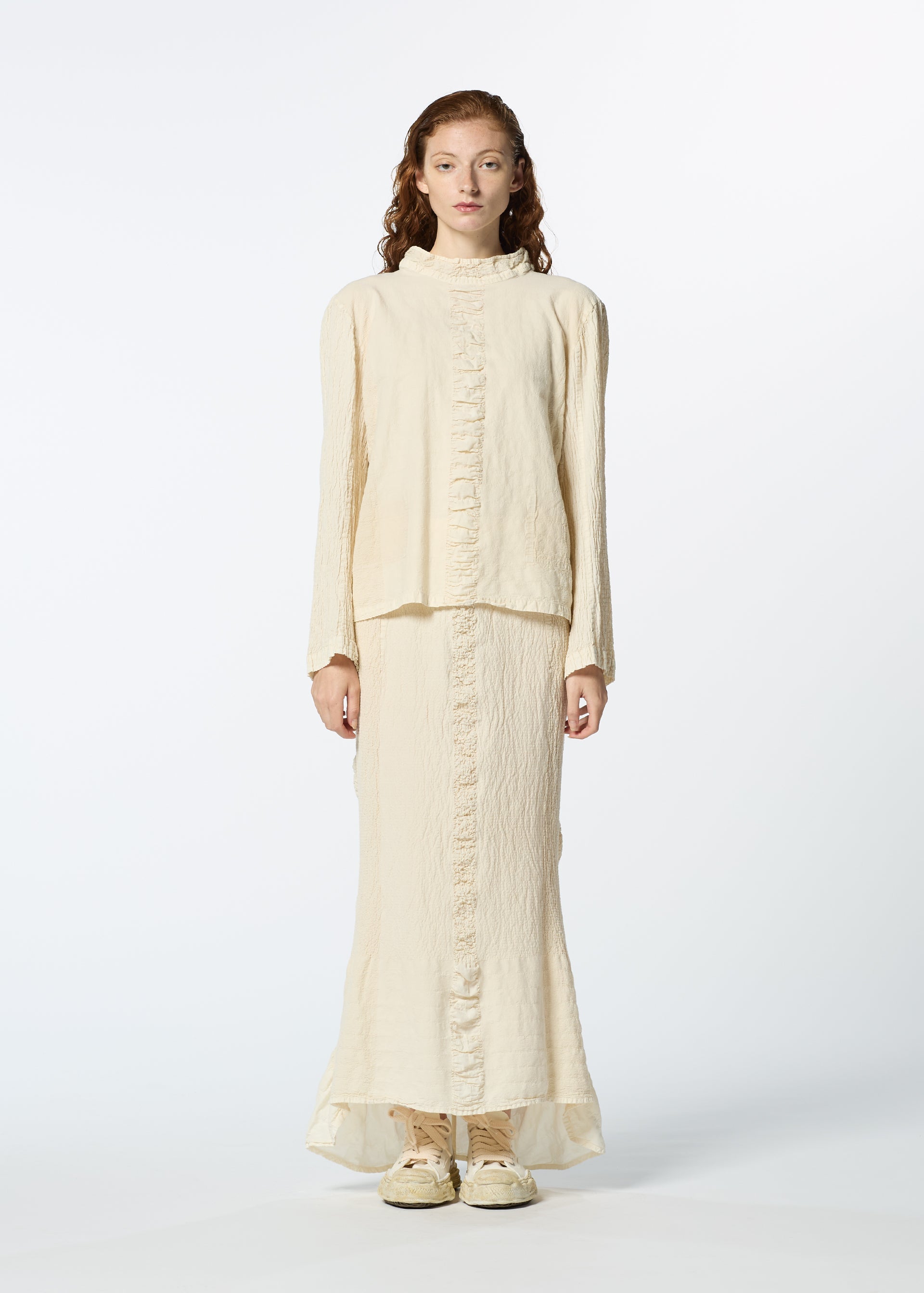 Zoomed view A woman with long, wavy hair wears HaaT's TENMOKU COTTON cream-colored, garment-dyed long-sleeved top and matching ankle-length skirt with subtle front ruffles, paired with beige shoes, standing against a white background.