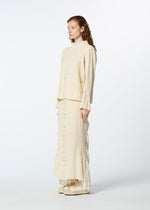A woman with long, wavy hair stands sideways against a white background, wearing HaaT’s TENMOKU COTTON loose-fit, ruffled long-sleeve top and matching skirt made from garment-dyed fabric, paired with cream shoes.