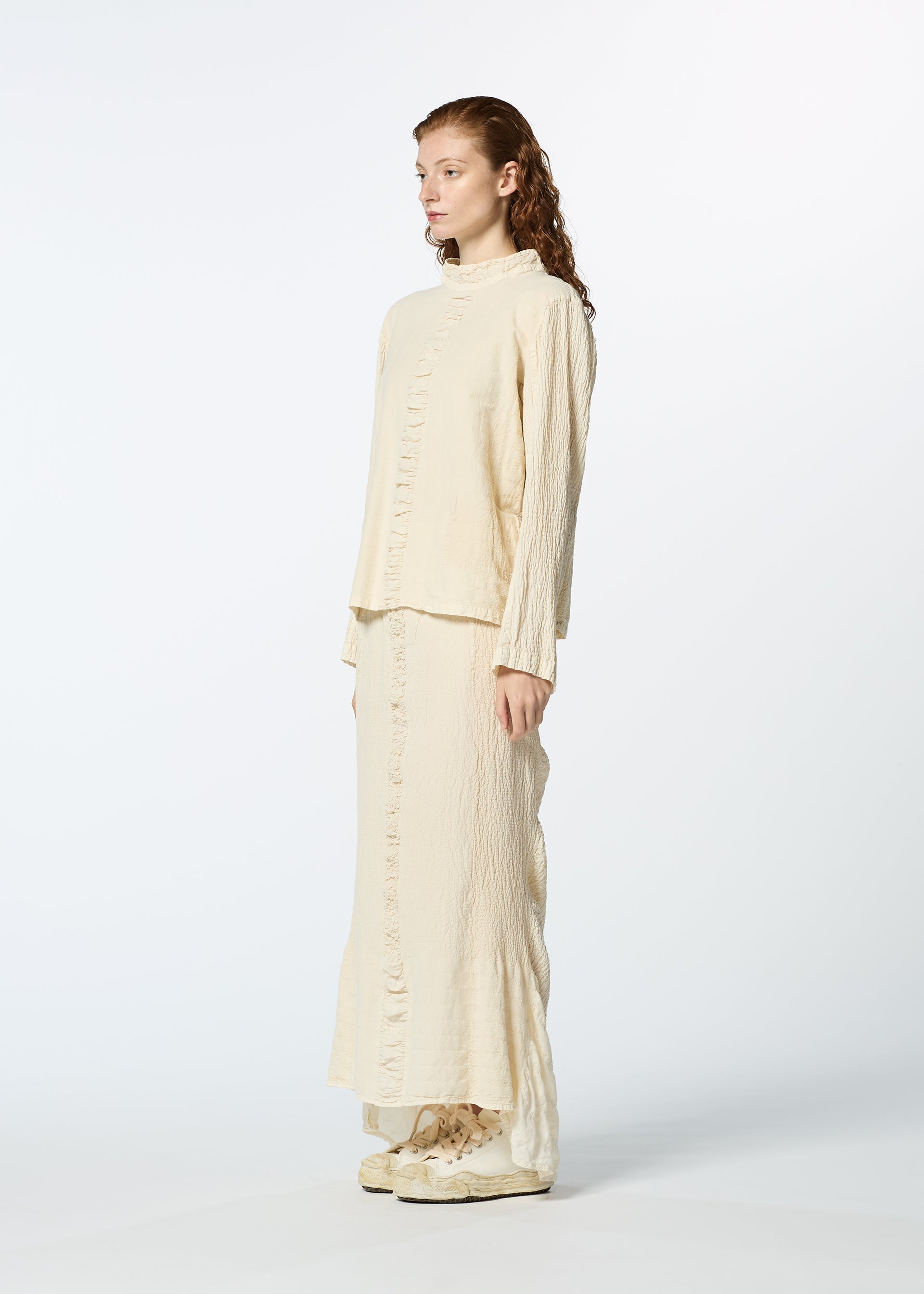 Zoomed view A woman with long, wavy hair stands sideways against a white background, wearing HaaT’s TENMOKU COTTON loose-fit, ruffled long-sleeve top and matching skirt made from garment-dyed fabric, paired with cream shoes.