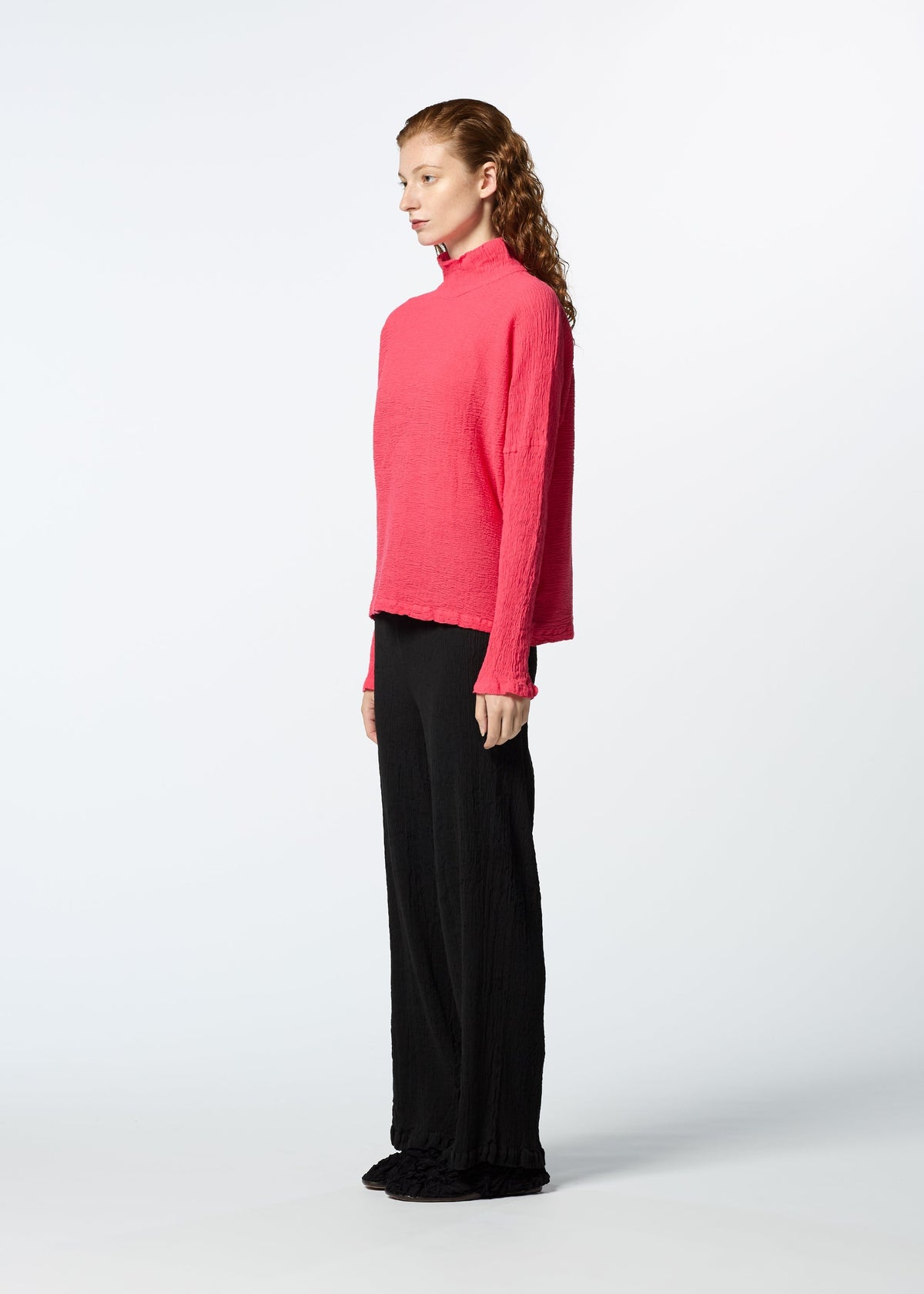 A person with long, wavy hair stands in profile against a plain white background, wearing the HaaT KYO CHIJIMI 2WAY JANUARY 2 vibrant pink high-neck long-sleeved top and wide-leg black pants. view 5 of 7