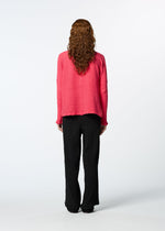 A person with long curly hair, dressed in the HaaT KYO CHIJIMI 2WAY JANUARY 2 bright pink high-neck top and black loose pants, stands facing away against a plain white background.