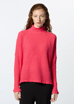A person with long, wavy red hair models the HaaT KYO CHIJIMI 2WAY JANUARY 2 bright pink high-neck long-sleeved top and black pants, standing against a plain white background and facing the camera.