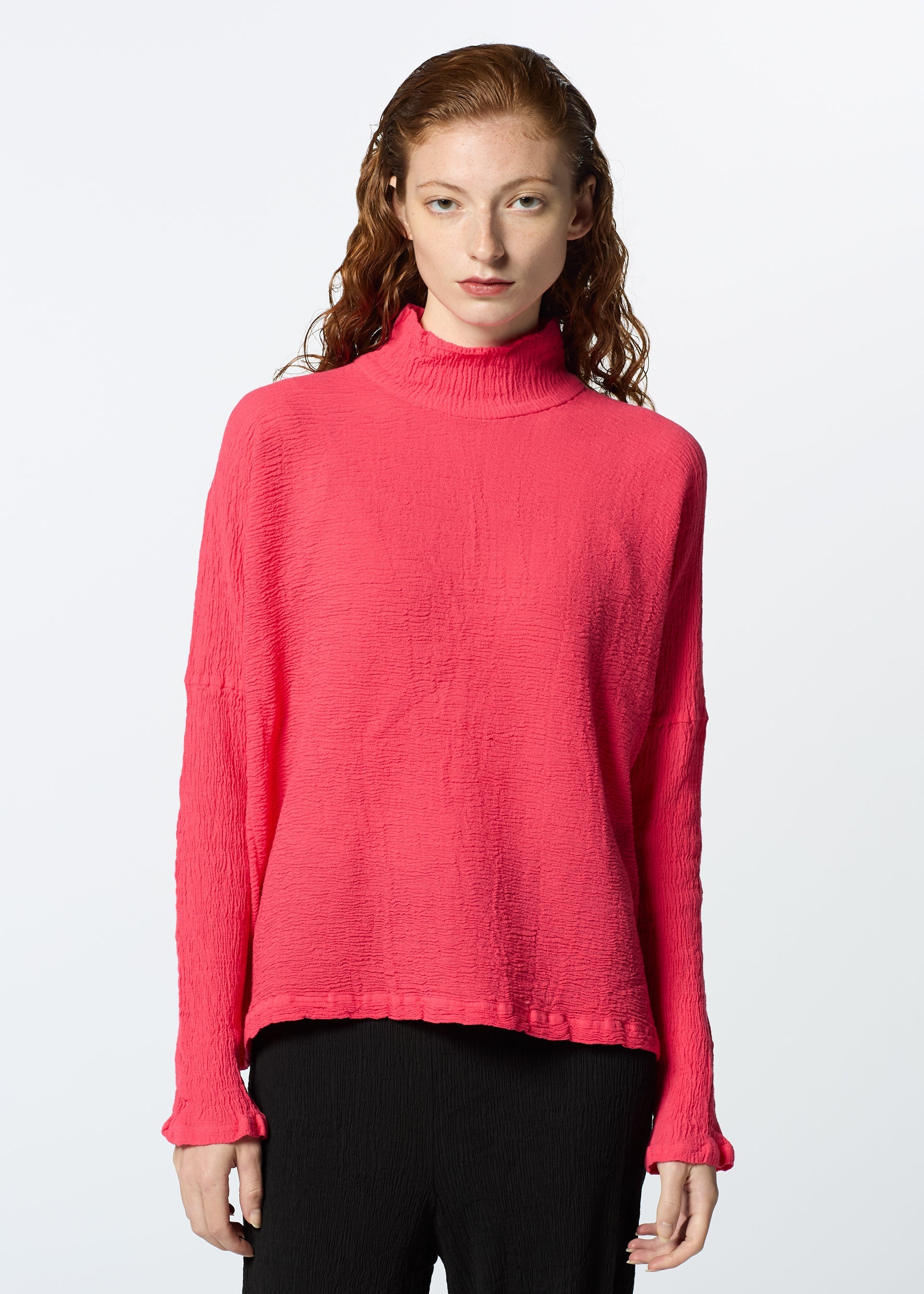 Zoomed view A person with long, wavy red hair models the HaaT KYO CHIJIMI 2WAY JANUARY 2 bright pink high-neck long-sleeved top and black pants, standing against a plain white background and facing the camera.