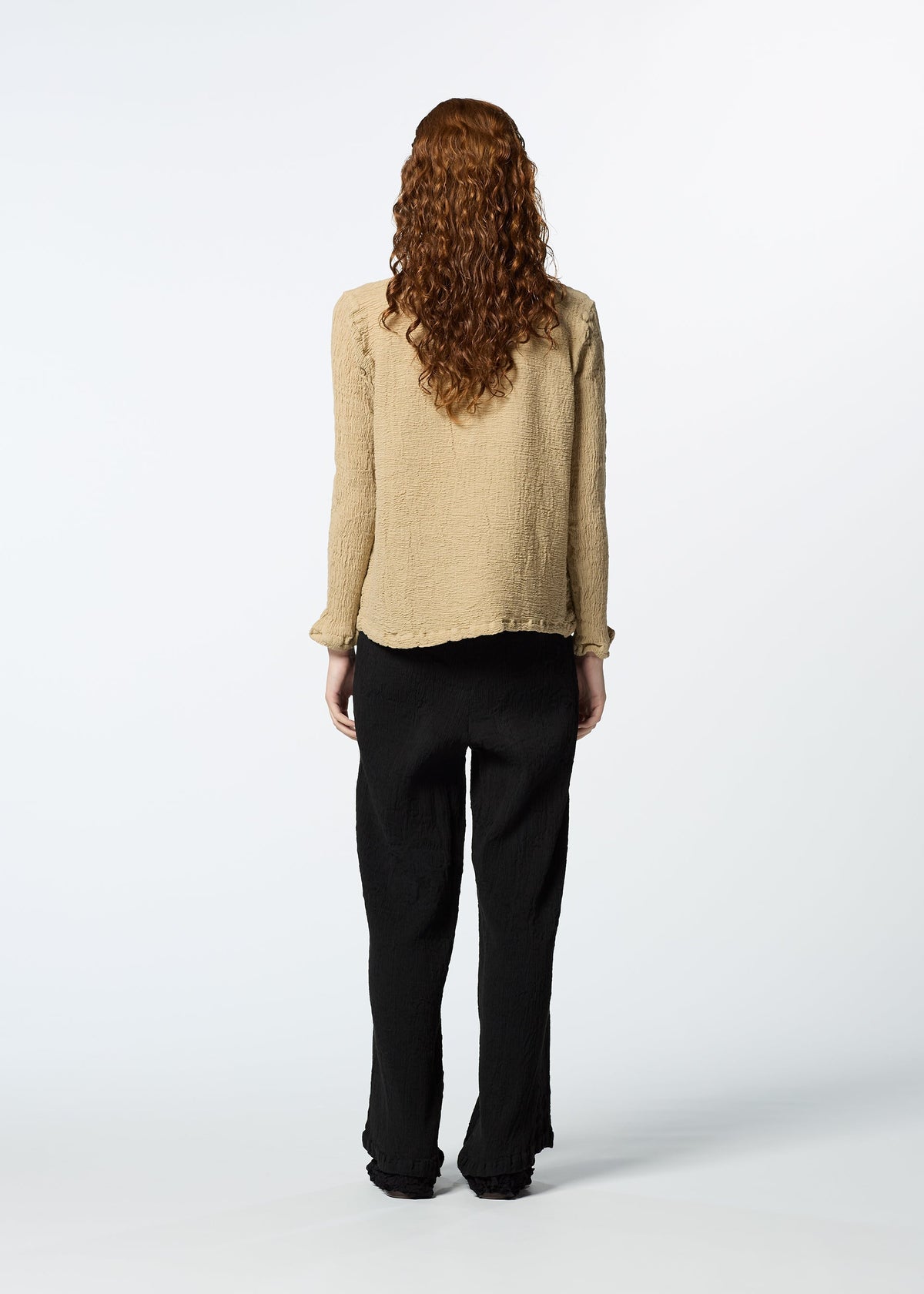 A person with long curly hair, dressed in HaaT’s KYO CHIJIMI 2WAY JANUARY 2 textured beige shirt and loose black pants, stands facing away against a plain white background. view 6 of 7