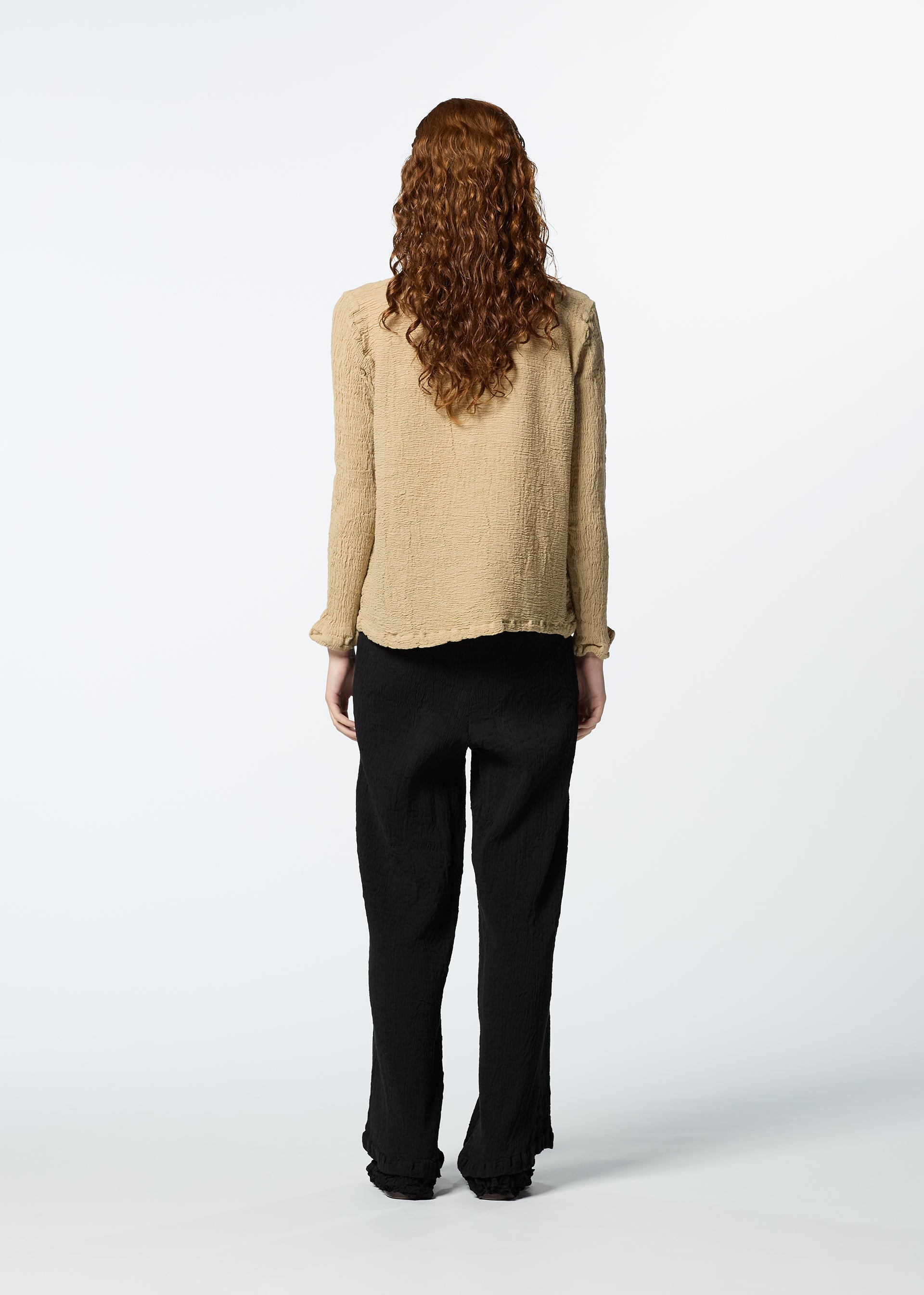 Zoomed view A person with long curly hair, dressed in HaaT’s KYO CHIJIMI 2WAY JANUARY 2 textured beige shirt and loose black pants, stands facing away against a plain white background.