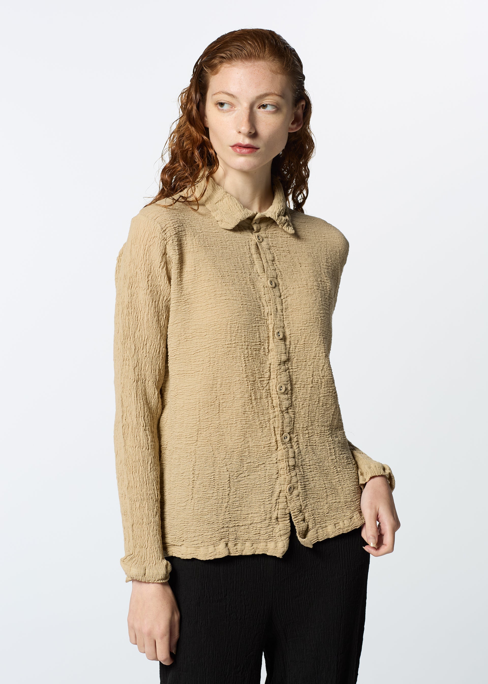 Zoomed view A person with wavy, reddish hair wears a beige HaaT KYO CHIJIMI 2WAY JANUARY 2 long-sleeved shirt made from hard-twist yarn, paired with black pants, standing against a plain light background.