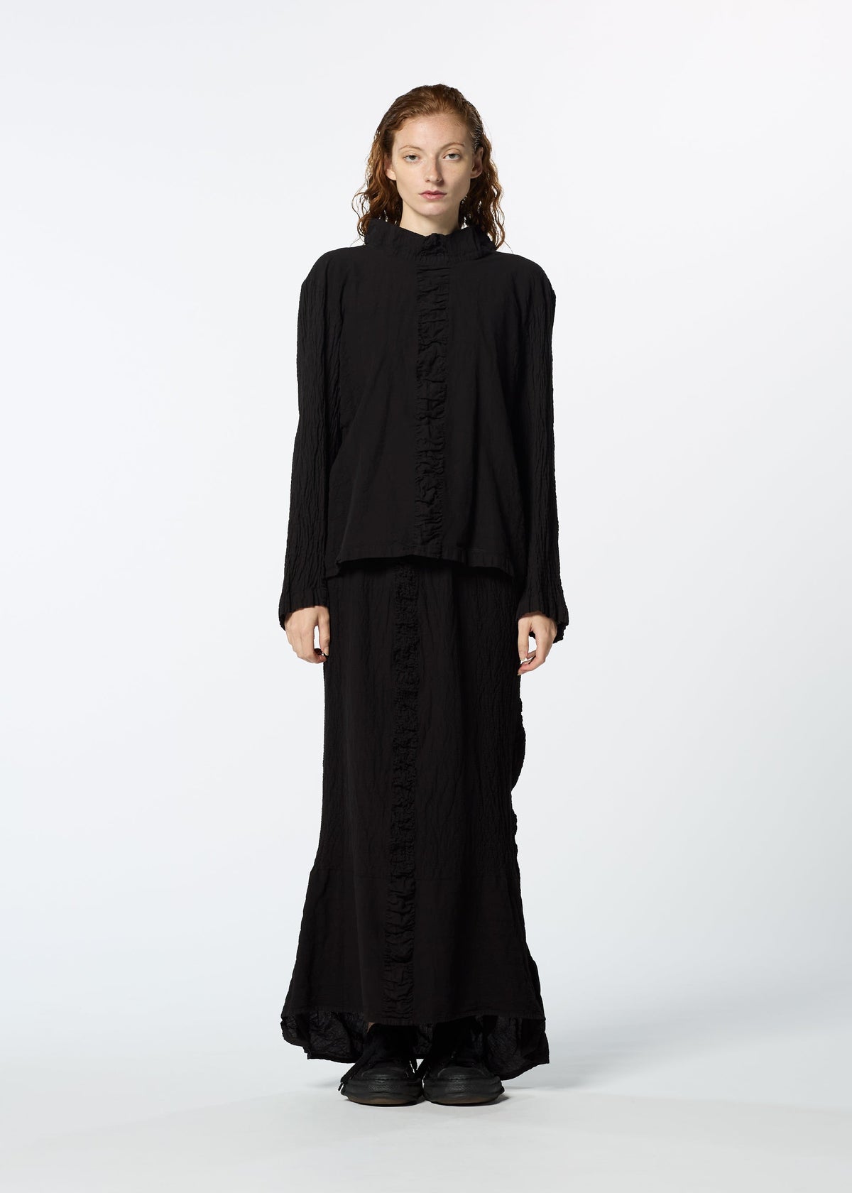 A person faces forward in a HaaT TENMOKU COTTON garment-dyed black long-sleeve top and matching long skirt with subtle ruffles, both made from textured stretchy fabric, paired with black shoes, against a plain white background. view 4 of 7
