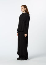 A person with long, wavy red hair stands sideways against a plain white background, wearing HaaT's TENMOKU COTTON garment-dyed black long-sleeve top and matching ankle-length skirt in textured stretchy fabric, paired with black shoes.