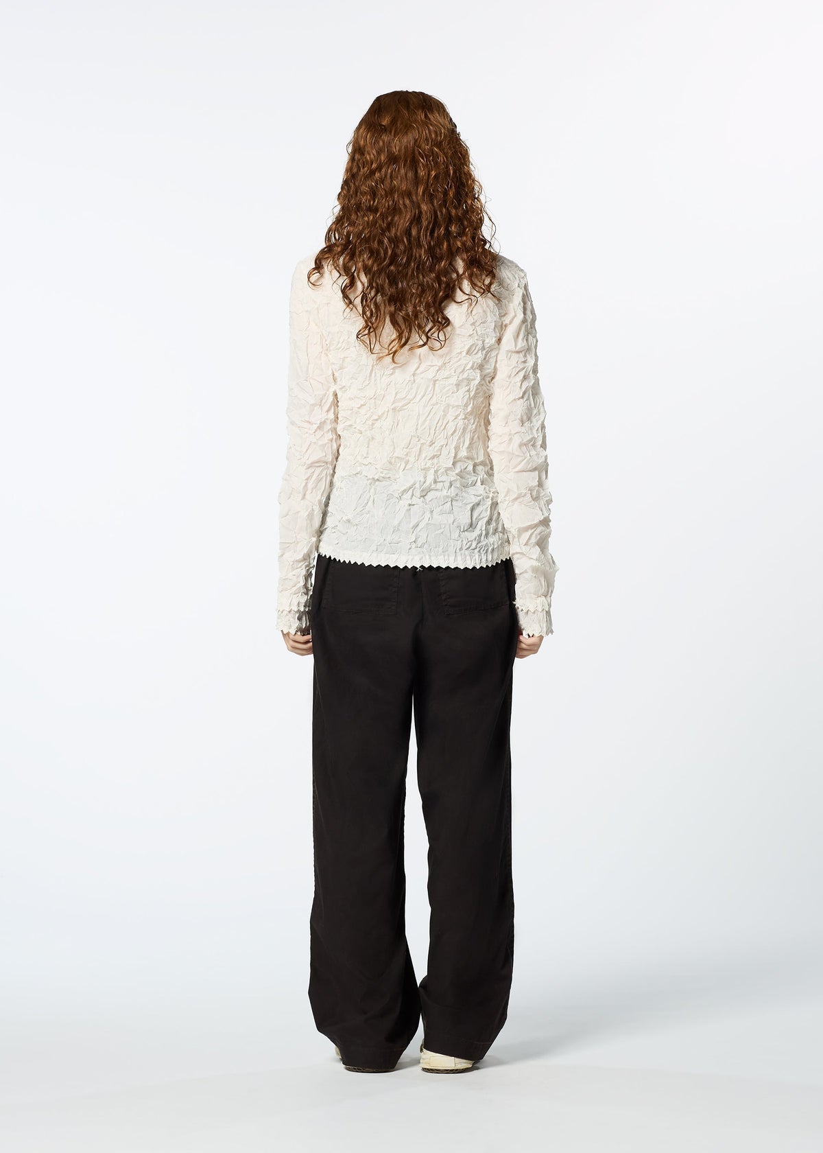 A person with long, curly brown hair stands facing away, wearing the HaaT KUMO SHIBORI SHIRT—a textured white long-sleeve piece—layered under a loose black cardigan and relaxed black pants against a plain white background. view 9 of 9