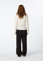 A person with long, curly brown hair stands facing away, wearing the HaaT KUMO SHIBORI SHIRT—a textured white long-sleeve piece—layered under a loose black cardigan and relaxed black pants against a plain white background.
