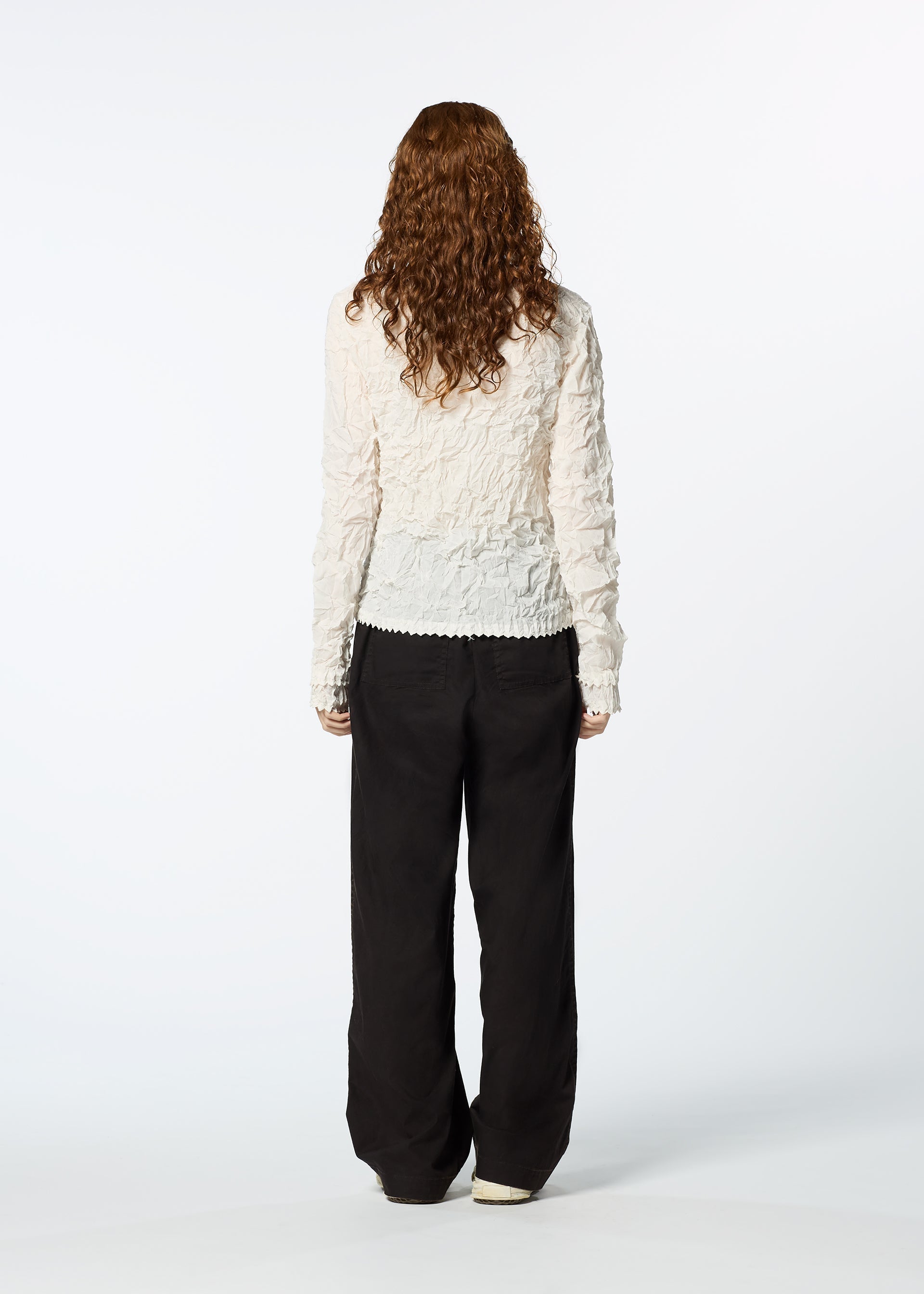 Zoomed view A person with long, curly brown hair stands facing away, wearing the HaaT KUMO SHIBORI SHIRT—a textured white long-sleeve piece—layered under a loose black cardigan and relaxed black pants against a plain white background.