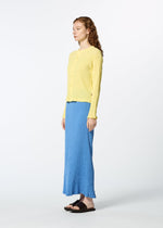 A woman with long curly hair stands in profile against a plain white background, wearing the HaaT KYO CHIJIMI MAY pale yellow round-neck long-sleeved cardigan made from ultra-fine cotton yarn, a blue ankle-length skirt, and black sandals.