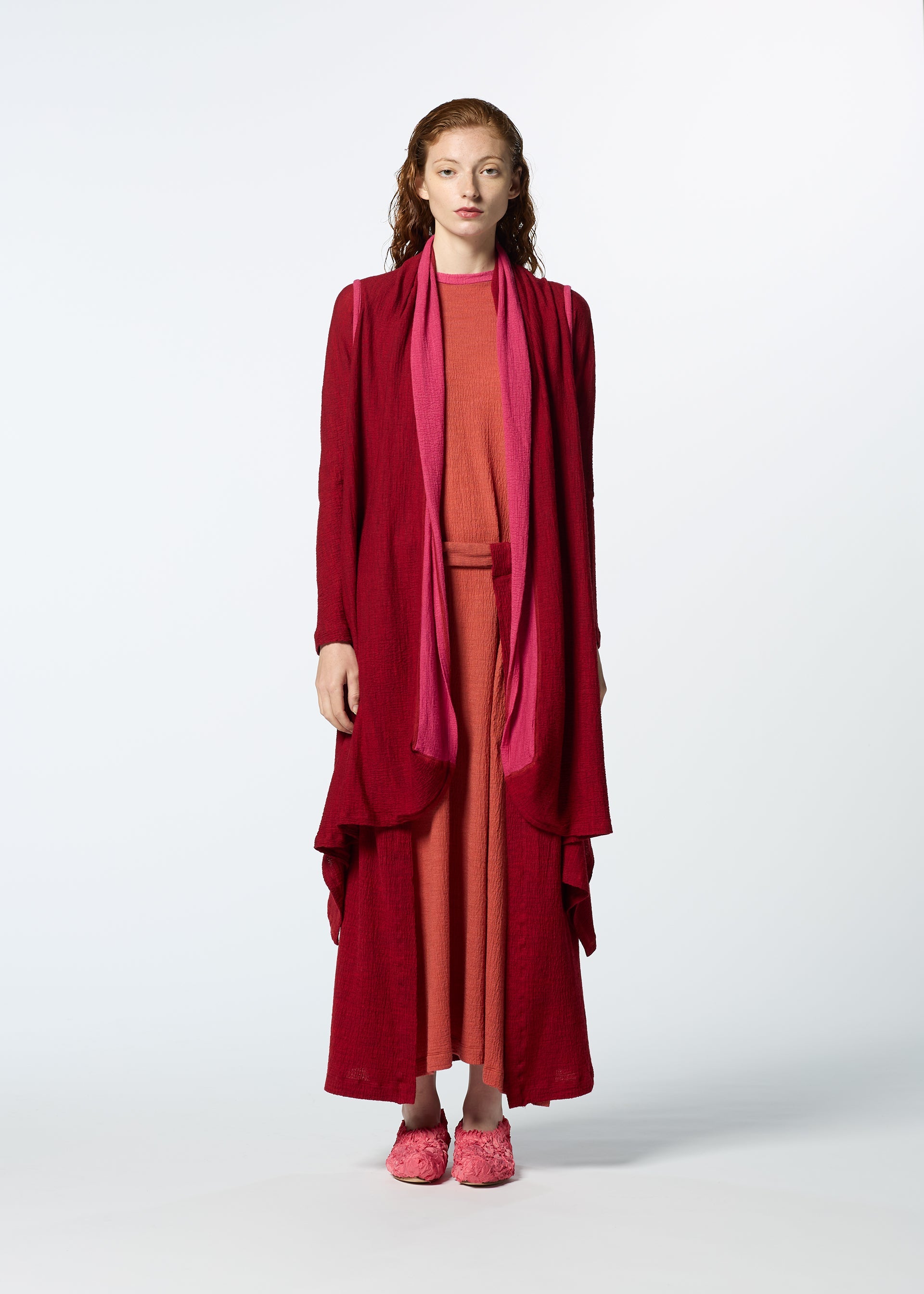 Zoomed view A woman faces forward in HaaT’s TRI COLOR WOOL: a long rose-pink dress with a flowing burgundy merino wool cardigan. She has long wavy hair and pink shoes, set against a plain light background.