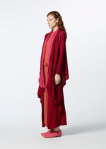 A woman with wavy brown hair stands in profile, wearing HaaT’s TRI COLOR WOOL layered red and pink outfit with matching pink slippers, against a plain white background.