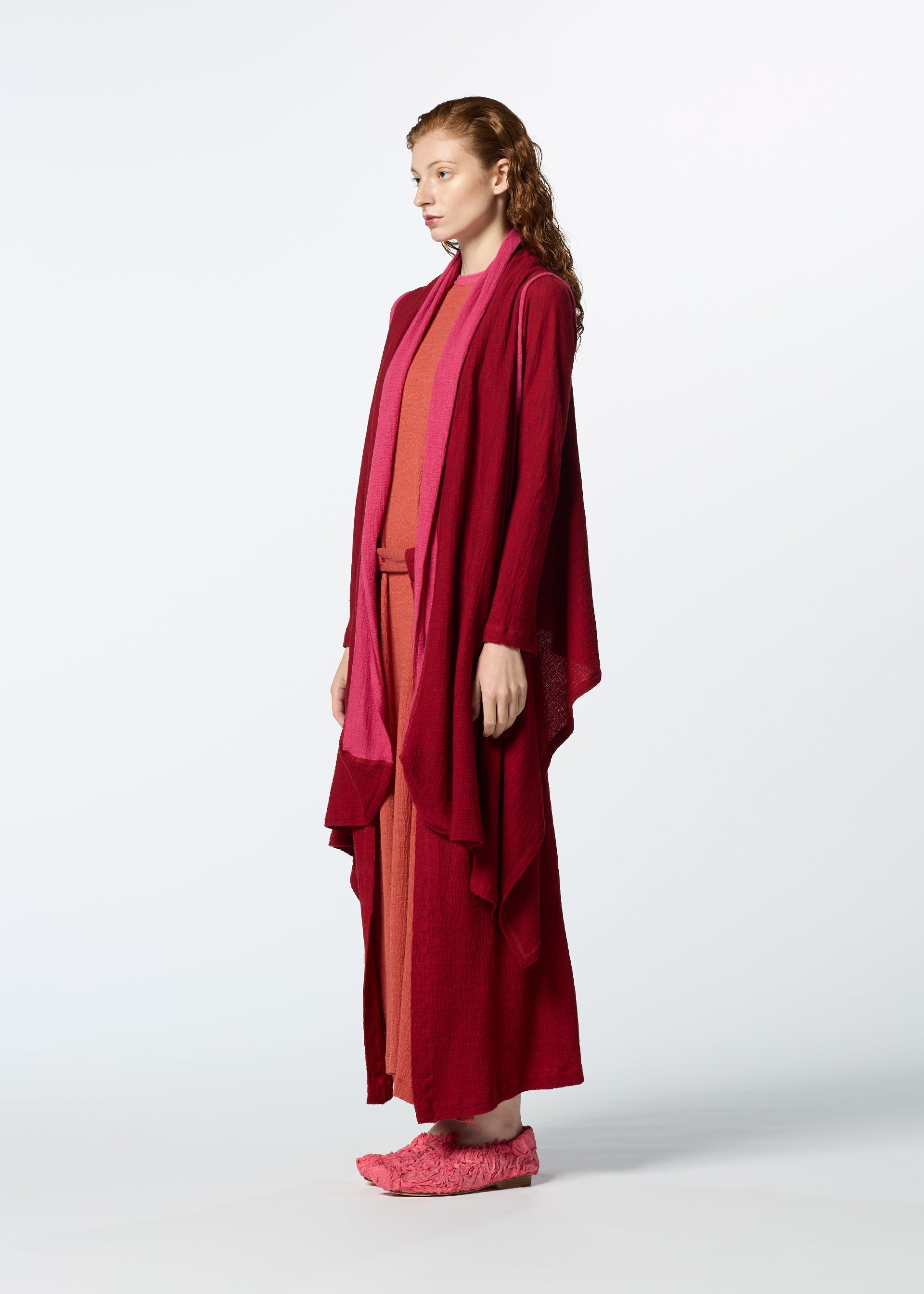 Zoomed view A woman with wavy brown hair stands in profile, wearing HaaT’s TRI COLOR WOOL layered red and pink outfit with matching pink slippers, against a plain white background.