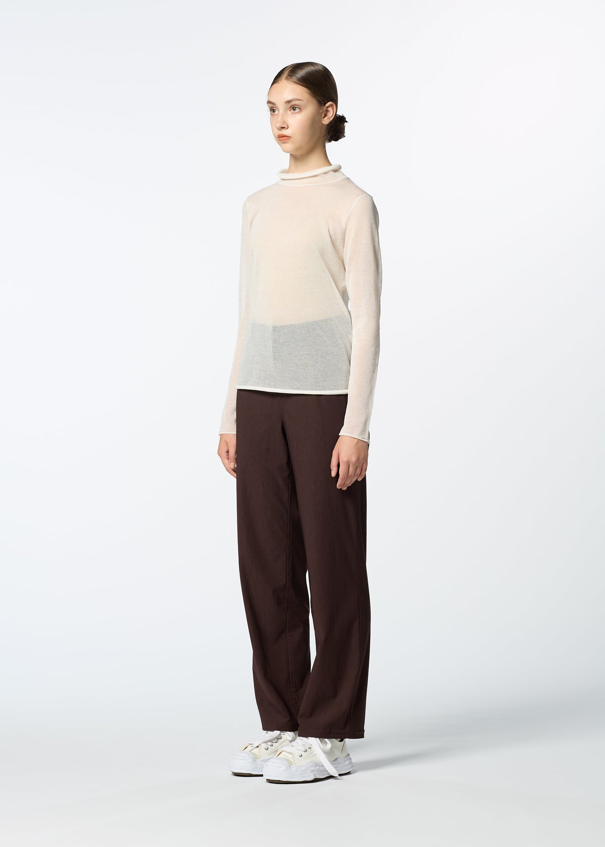 A person with hair pulled back stands against a plain background, wearing HaaT’s NYLON STRETCH BOTTOM in dark brown, a sheer beige long-sleeve top, and white sneakers while gazing slightly to the side. view 7 of 8