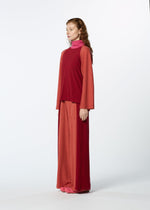 A woman with long wavy hair stands in profile, wearing HaaT's TRI COLOR WOOL relaxed fit, long-sleeved red top and merino wool pants with a pink turtleneck and pink shoes against a plain white background.