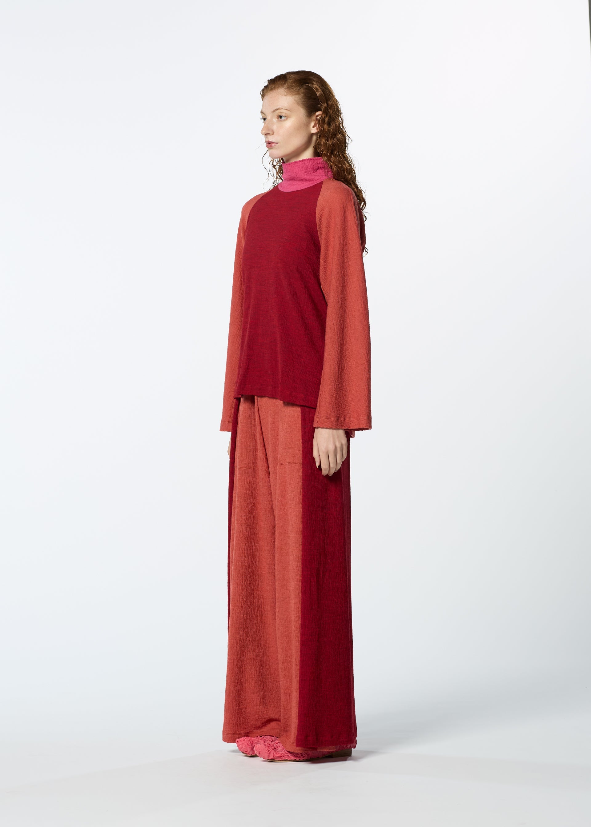 Zoomed view A woman with long wavy hair stands in profile, wearing HaaT's TRI COLOR WOOL relaxed fit, long-sleeved red top and merino wool pants with a pink turtleneck and pink shoes against a plain white background.