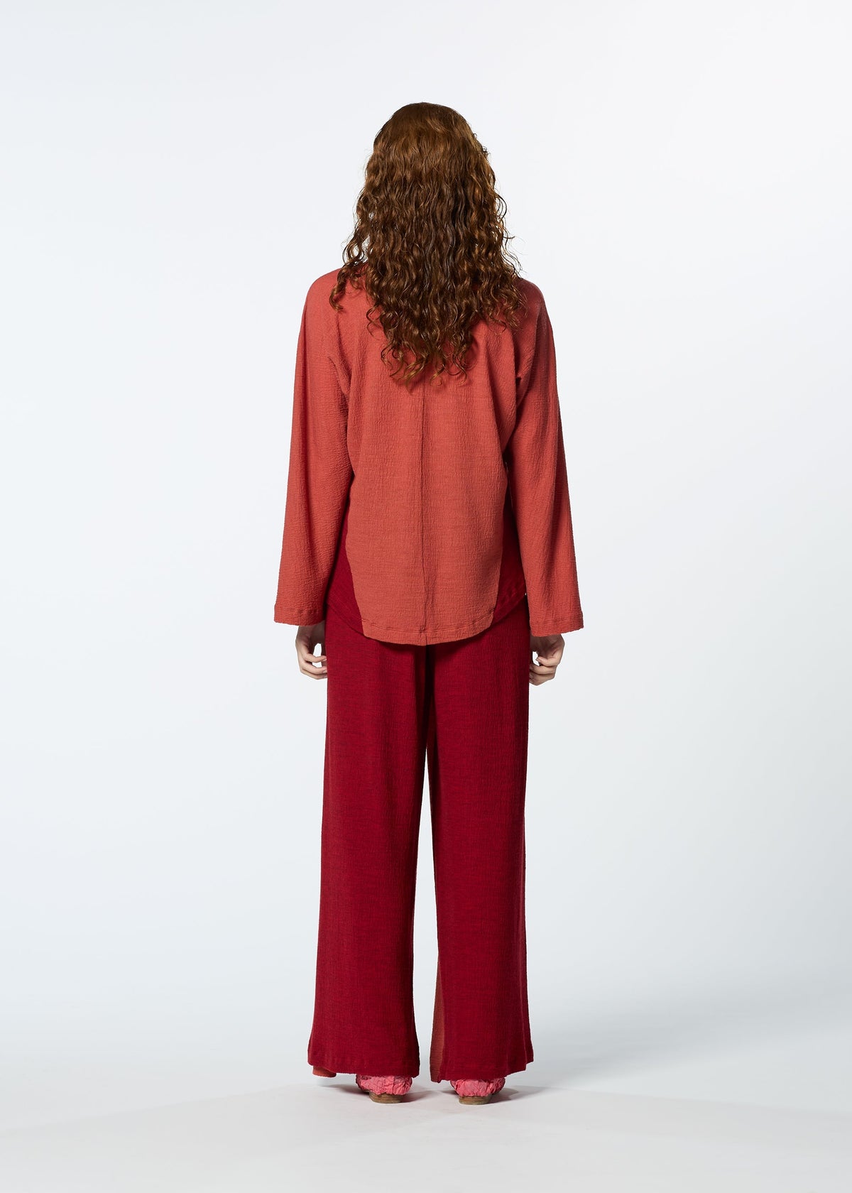 A person with long, curly brown hair faces away, wearing HaaT's TRI COLOR WOOL loose, long-sleeve reddish top and relaxed fit maroon merino wool pants against a plain white background. view 12 of 13