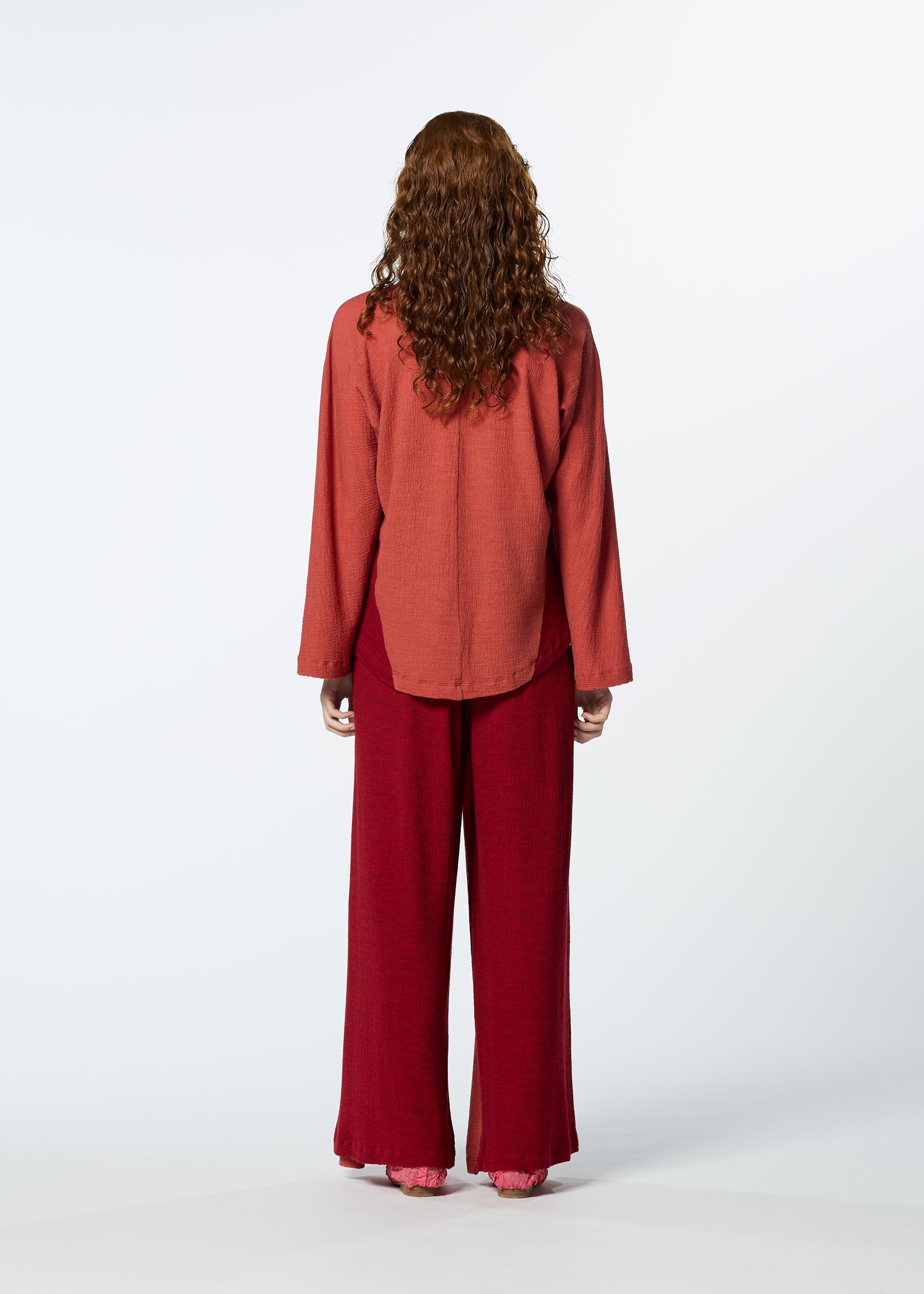 Zoomed view A person with long, curly brown hair faces away, wearing HaaT's TRI COLOR WOOL loose, long-sleeve reddish top and relaxed fit maroon merino wool pants against a plain white background.