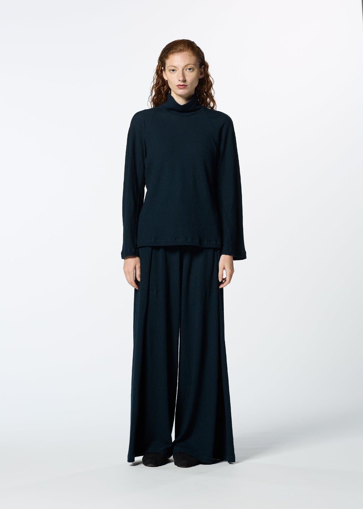 A person with long curly hair faces forward against a white background, wearing HaaT’s TRI COLOR WOOL black turtleneck sweater and relaxed fit merino wool pants. view 13 of 13