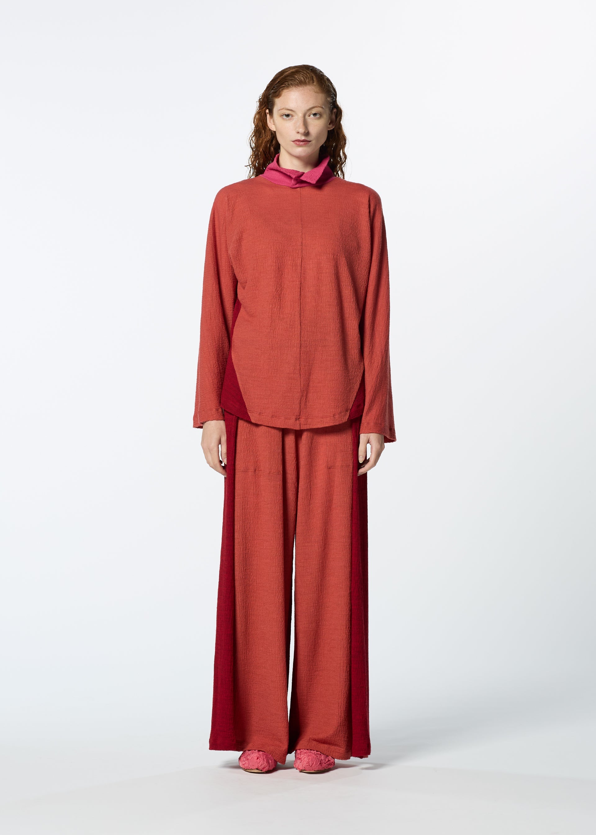 Zoomed view A person with wavy hair stands facing forward in HaaT's TRI COLOR WOOL: a loose-fitting, long-sleeve reddish-orange merino wool top and wide-leg matching pants, paired with flat shoes against a plain white background.