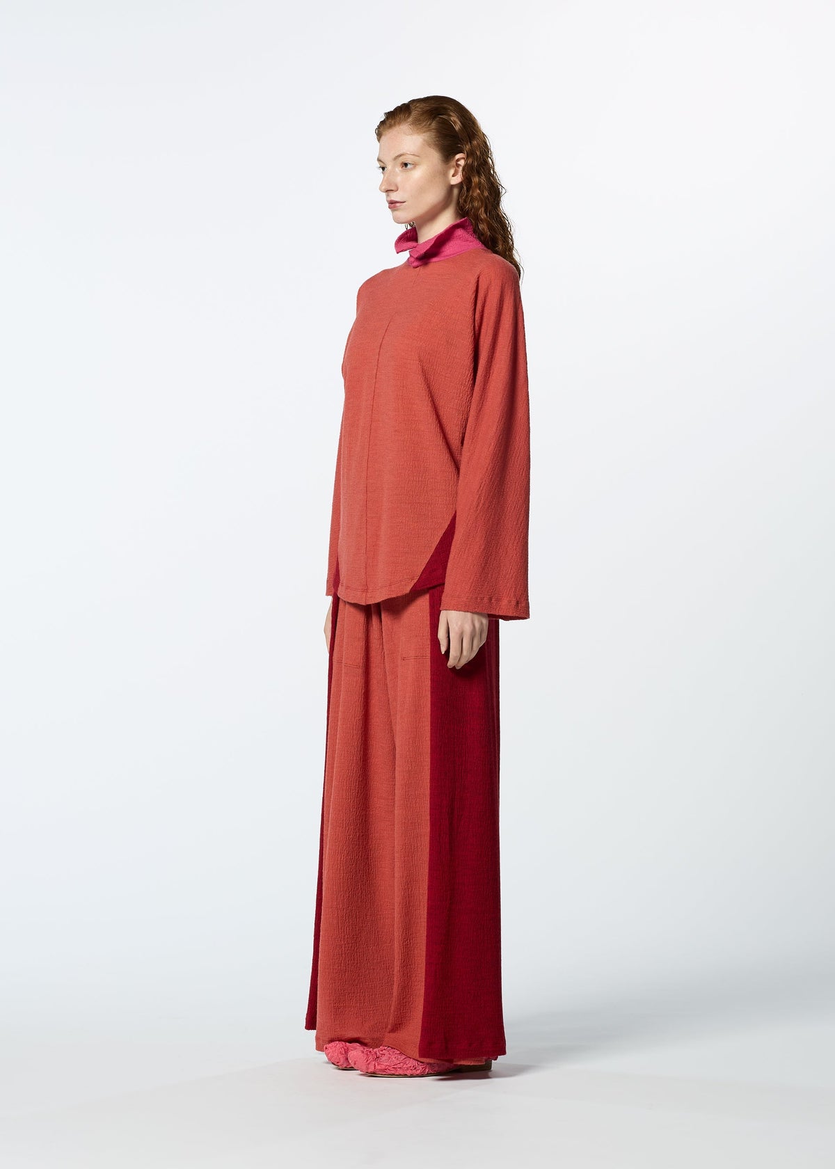 A woman with red hair stands sideways against a white background, wearing the HaaT TRI COLOR WOOL set: a loose, long-sleeved reddish-orange top with a pink collar, matching wide-leg pants, and pink shoes. view 14 of 16