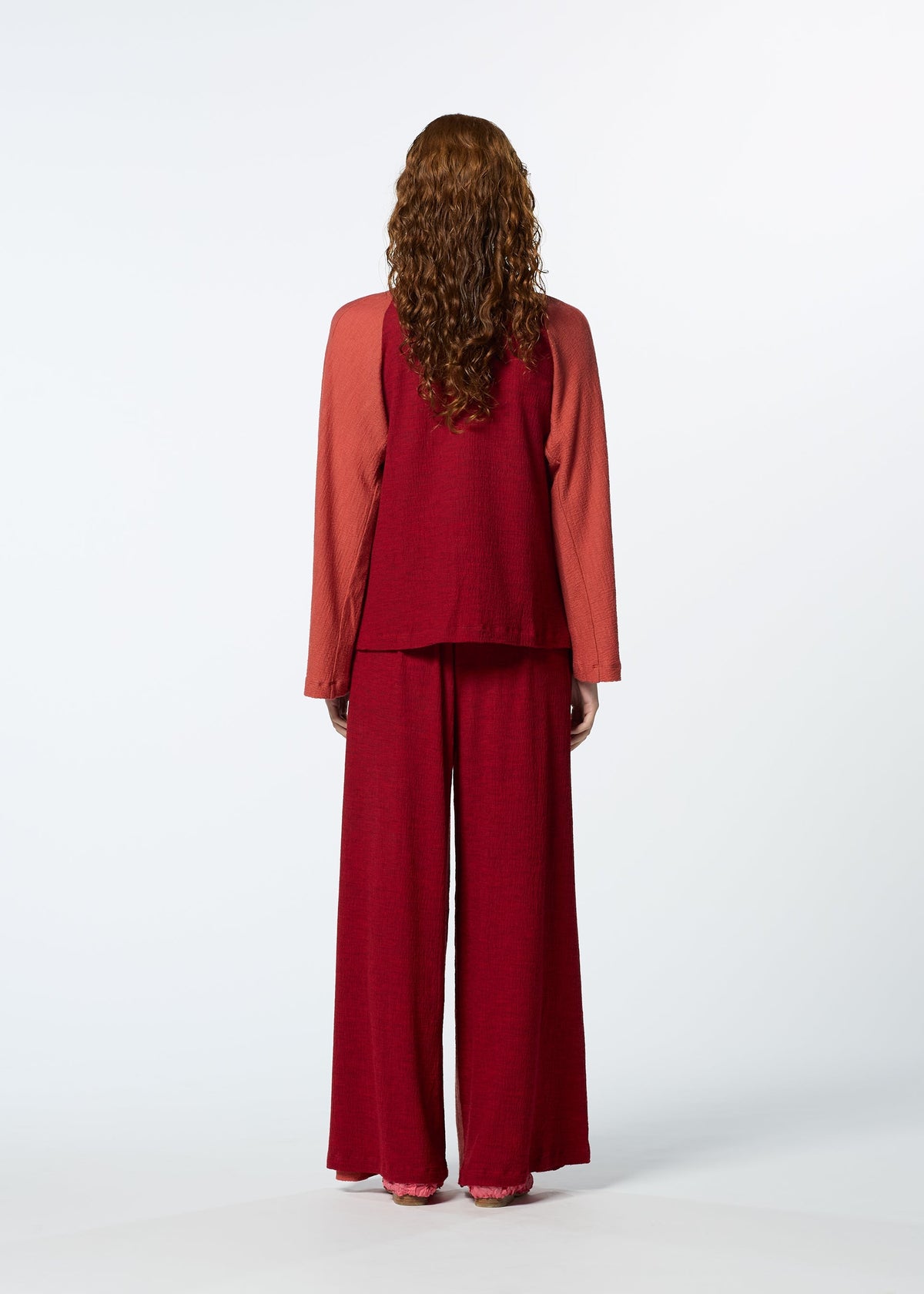 A person with long curly hair faces away, wearing HaaT's TRI COLOR WOOL loose-fitting, garment-dyed reddish long-sleeved top and matching wide-leg pants against a plain light background. view 15 of 16