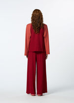 A person with long curly hair faces away, wearing HaaT's TRI COLOR WOOL loose-fitting, garment-dyed reddish long-sleeved top and matching wide-leg pants against a plain light background.