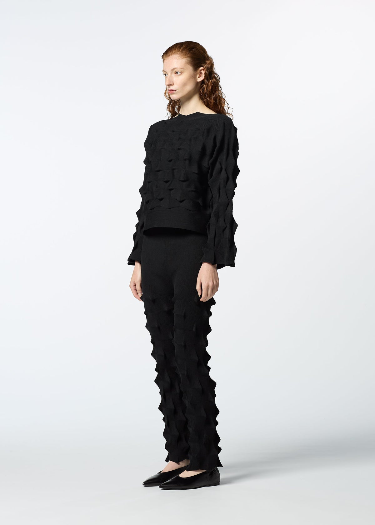 A woman with long, wavy hair stands against a white background wearing HaaT’s ZIG ZAG KNIT black pants and a textured organic cotton and recycled polyester sweater, paired with black flats. She gazes off to the side. view 6 of 7