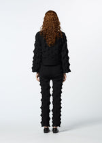 A person with long, curly hair in a textured black outfit faces away, showcasing HaaT’s ZIG ZAG KNIT pants made from organic cotton and recycled polyester with geometric raised patterns, paired with black shoes against a white background.
