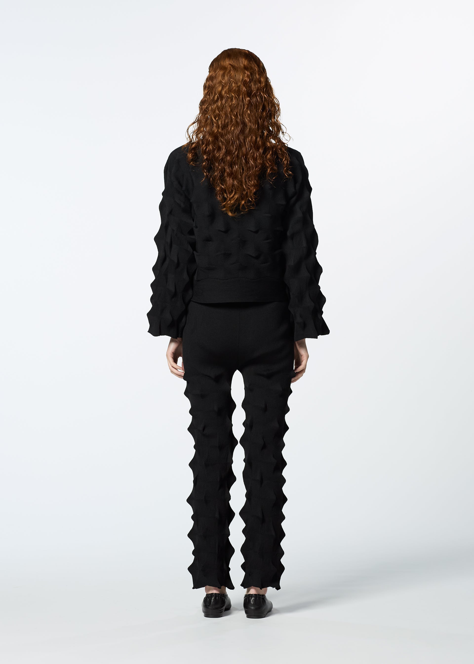 Zoomed view A person with long, curly hair in a textured black outfit faces away, showcasing HaaT’s ZIG ZAG KNIT pants made from organic cotton and recycled polyester with geometric raised patterns, paired with black shoes against a white background.