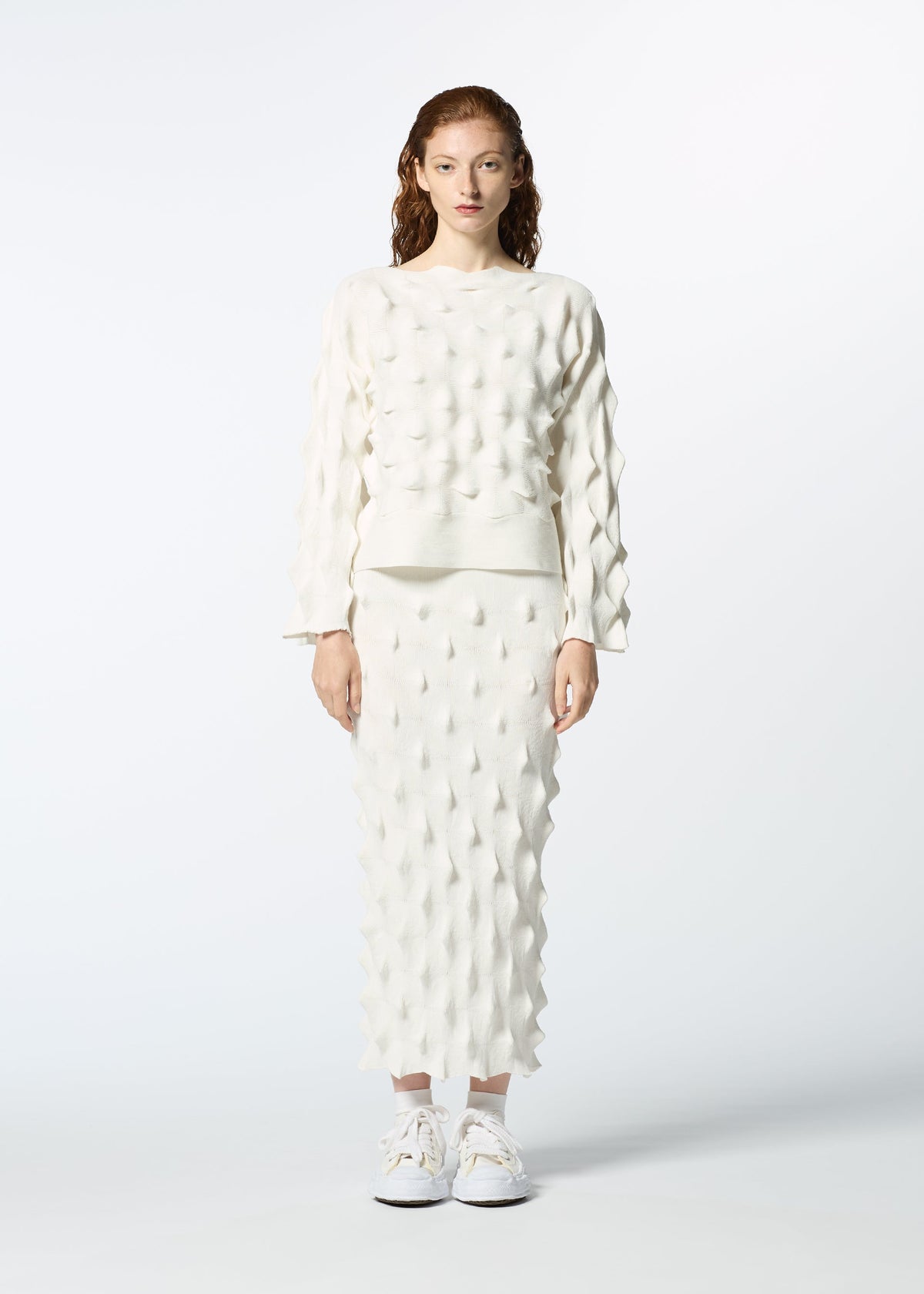 A person faces forward in HaaT’s ZIG ZAG KNIT set—white textured sweater and zigzag knit skirt crafted from organic cotton and recycled polyester—paired with white sneakers, against a plain white background. view 5 of 7