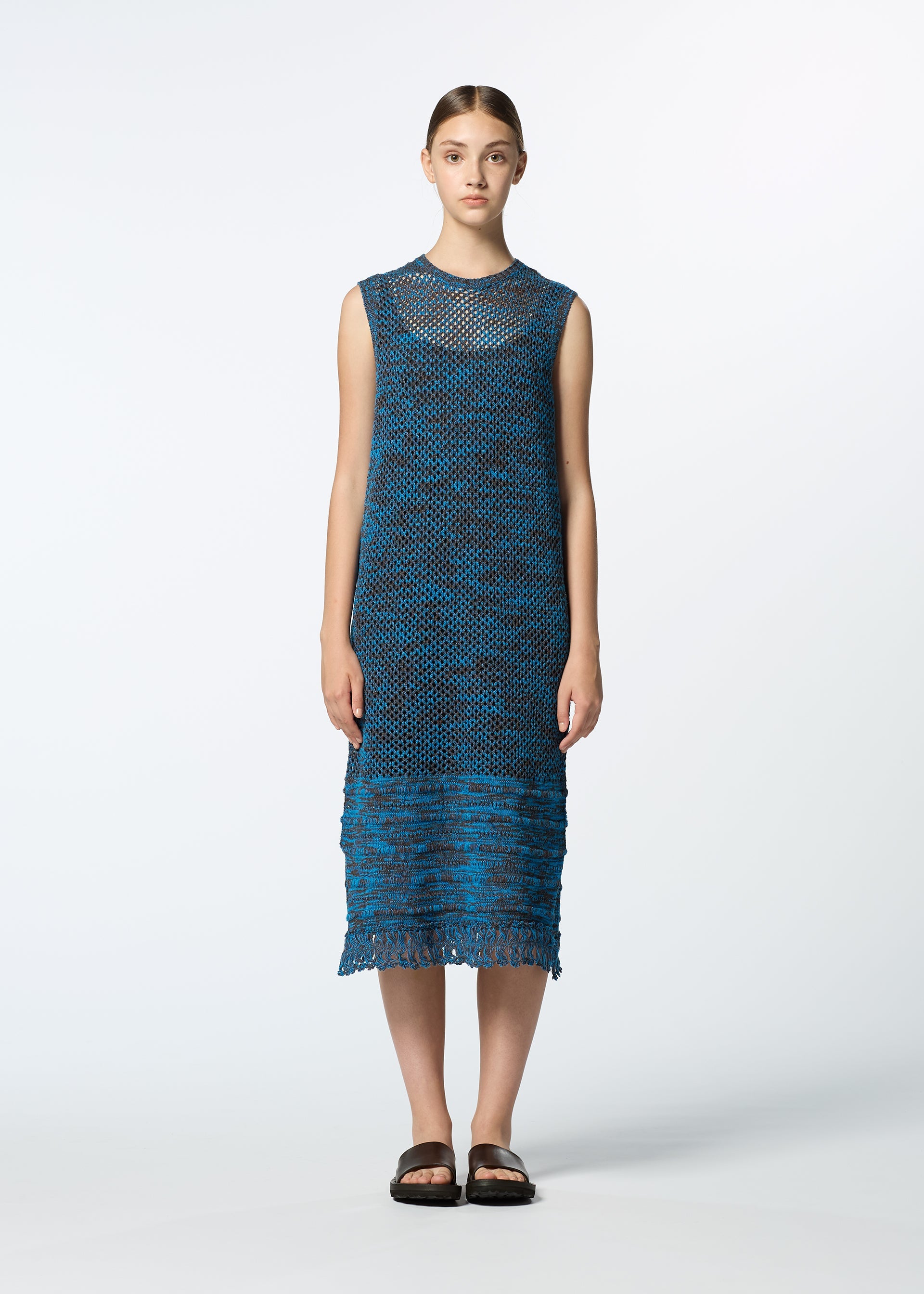 Zoomed view A young woman with straight brown hair stands facing forward, wearing HaaT's MELANGE MESH sleeveless blue knit dress with a textured pattern and fringe hem, paired with black open-toe sandals against a plain white background.